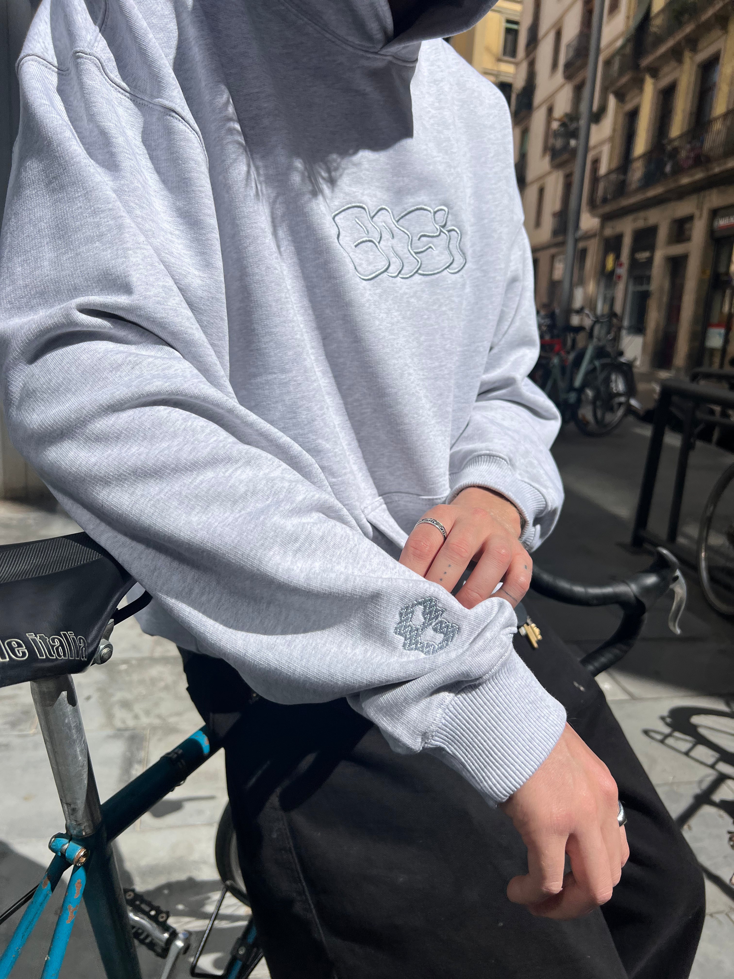 Person wearing a light gray sweatshirt with a logo, sitting on a bicycle in an urban setting.